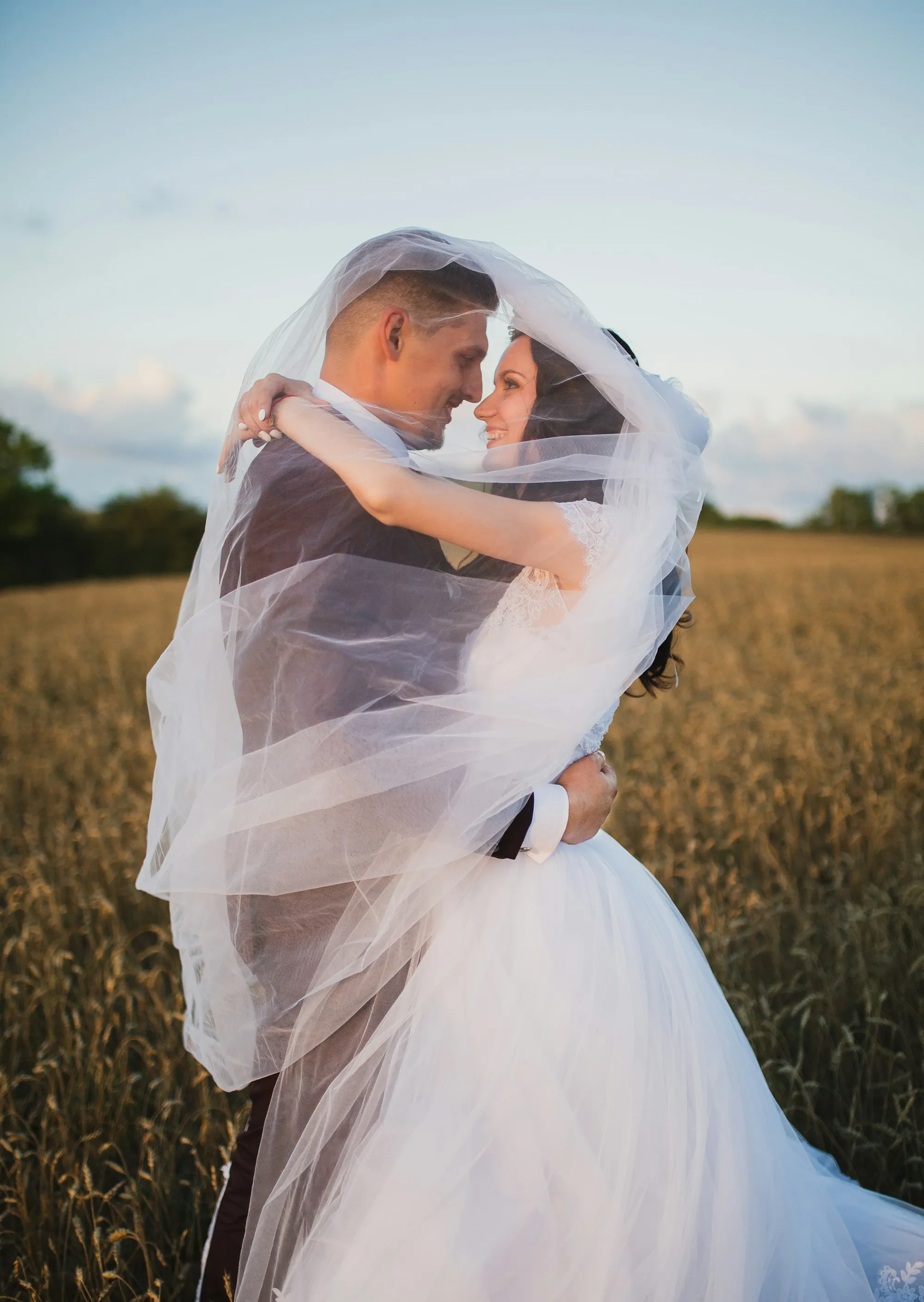 A bride and groom wrapped together in her veil in a golden field