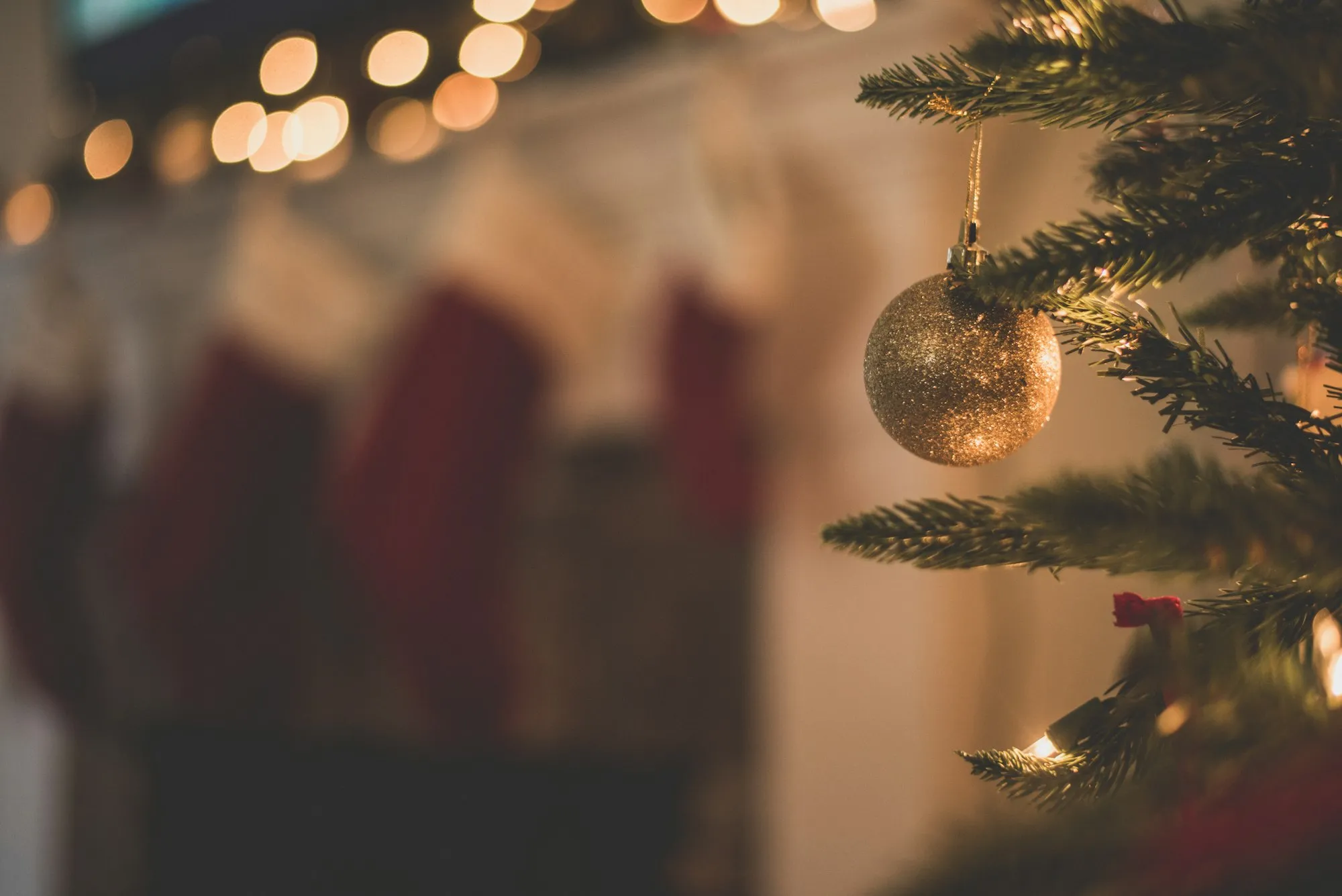 A gold ornament on a Christmas tree with stockings glowing behind