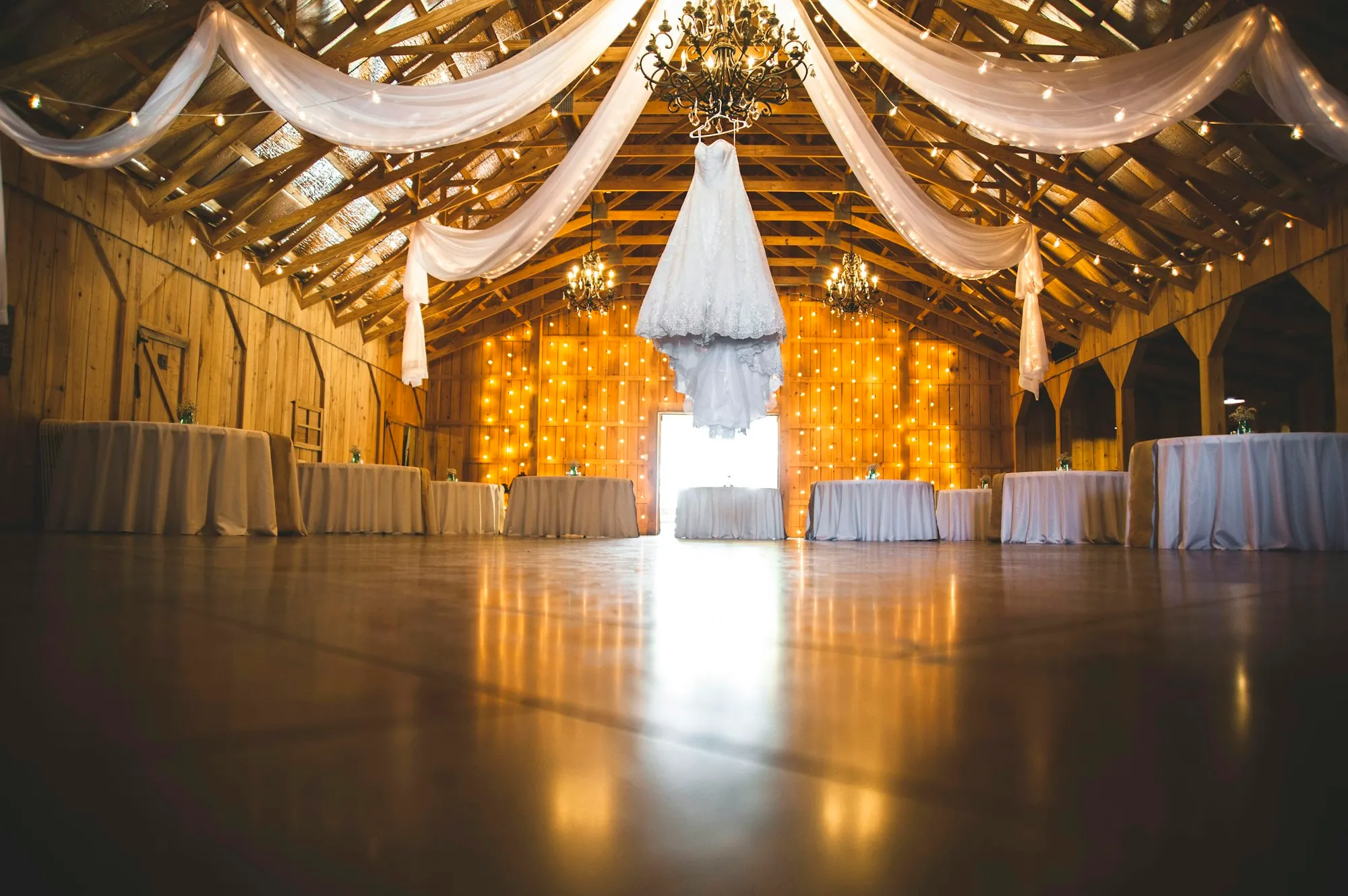 A barn reception with draped fabric, a chandelier and a wall of warm string lights